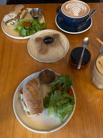 A table is set with culinary delights. A blue mug of latte art sits beside a small plate of pastries. A jar of chili oil and a container of sauce are nearby. Two plates hold sandwiches with green salad and small metal cups of dressing. The sandwiches appear to be a hearty, crusty variety.