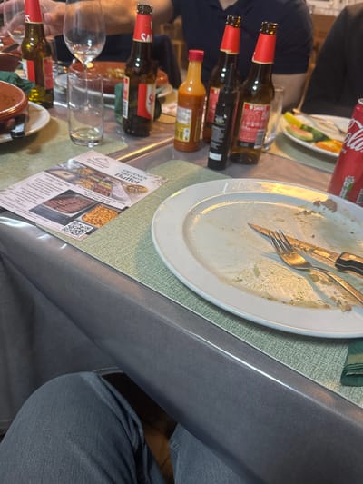 The image depicts an indoor dining scene at the Restaurant Bahnhof Lalden in Lalden, Switzerland. A grey, patterned plastic tablecloth covers a table, overlaid with green placemats. In the foreground, an oval white plate, largely cleared of food, holds a fork and a knife with a black handle. To the left of the plate, a flyer for an "Offenes Mittags Buffet" from the "Restaurant Bahnhof Lalden" is displayed, featuring images of food items and a QR code.

Multiple beverage containers are distributed across the table: at least four brown glass bottles with red "Sagres" labels, an orange bottle of sauce, a dark bottle, an empty clear glass tumbler, a clear wine glass, and a red Coca-Cola can. A partially visible plate of fruit, including watermelon and orange slices, is present in the background right. At least three individuals are partially visible at the table. The lower legs of a person wearing grey trousers are seen in the immediate foreground. A person in a dark blue polo shirt is visible in the background, with an arm extended towards the table. The scene indicates a meal that has concluded or is in its final stages.