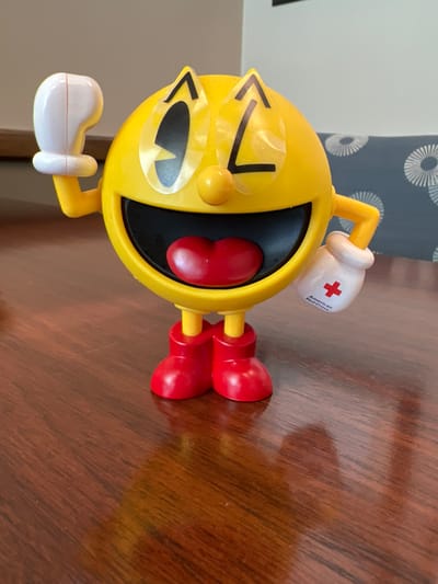 A yellow Pac-Man toy, with a white glove on its left hand and a white glove with a red cross and "American Red Cross" printed on it on its right hand, stands on a wooden table. The toy has a wide open mouth with a red heart-shaped tongue, black eyes with yellow pupils, and yellow feet. The background shows a blurry couch with a circular pattern.