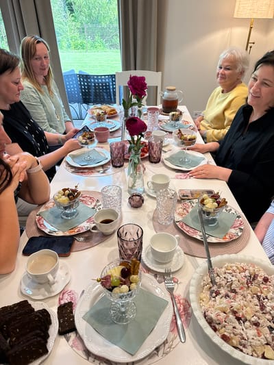 A group of seven women are seated around a table indoors for what appears to be a brunch or tea gathering. The table is set with floral-patterned placemats and plates, various dishes of food, and drinks. The women are casually dressed and engaged in conversation. One woman in the foreground is looking at her phone. The room has a large window with a view of greenery and what appears to be outdoor furniture. The setting suggests a home or a small cafe. The location is Rāmava, Latvia, as indicated by a watermark on the original image.