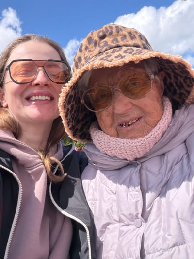 A photo taken from a low angle shows two individuals outdoors against a bright blue sky with scattered clouds, suggesting daylight. A younger woman with fair skin and shoulder-length brown hair is in the foreground wearing large square-framed sunglasses. She is smiling, revealing her teeth. She has on a pinkish hoodie with the zipper partially open and a black jacket underneath. Behind her, an older woman with wrinkled skin and a few visible gold teeth is wearing glasses with a pink, knitted cap and a leopard-print bucket hat. She also wears a light-colored, puffy coat. The setting is undefined, and the location in Valozhyn, Belarus, cannot be confirmed from visual elements.