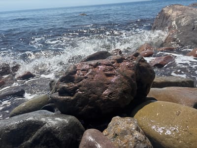 The image depicts a rocky shoreline with waves crashing against the rocks. The foreground features several large, wet rocks of varying colors and textures, including gray, brown, and tan. The dominant central rock has a dark, rugged surface with reddish-brown hues, and is covered in water droplets. In the background, the sea is visible with white foam and waves washing towards the shore. The sky is light blue and hazy. The location is likely a beach in Semidvorye, Ukraine, given the context. There are no people visible in the image.