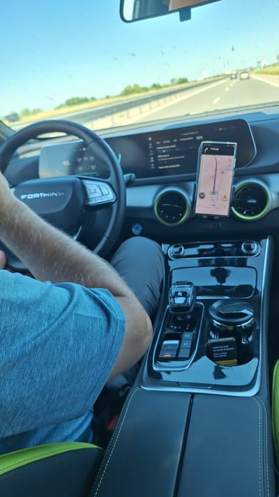 The image shows the interior of a moving car, specifically the driver's side. The car is traveling on a highway with green trees and vegetation visible in the background. The driver is wearing a blue shirt and gray pants, with their hands on the steering wheel. A mobile phone displaying a navigation app is mounted on the dashboard, showing a route with a distance of 118 km. The car's dashboard features a digital display and air vents with lime green accents. A black center console is visible with a gear shifter and control buttons. A card is visible in the console, and the text "Завинаги във времето" can be read on it. There is a rear-view mirror visible on the left side of the frame. No specific location in Opalchenets, Bulgaria can be determined from the image alone.