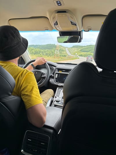 A person wearing a yellow t-shirt and a black bucket hat is driving a car. The car's interior is visible, with black leather seats and a dashboard featuring a central touchscreen display. The driver's hands are on the steering wheel. Outside the car, a winding road is visible, surrounded by green hills and trees under a cloudy sky. There are power line towers in the distance. The scene appears to be on a road in or near Krasnaya Gorka, Russia, given the description of the landscape.