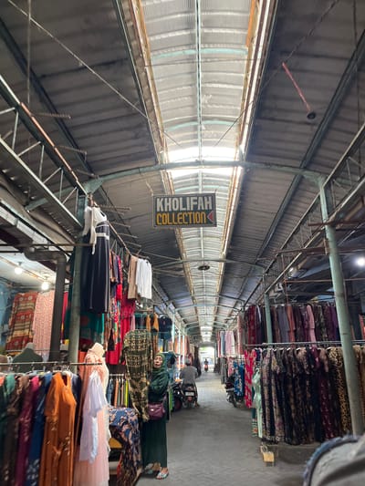 The image depicts a marketplace with stalls selling clothing, likely in Gedang, Indonesia. The marketplace features a long, covered walkway with a corrugated metal roof and exposed metal beams, allowing natural light to filter through. Rows of clothing racks line both sides of the walkway, displaying a variety of garments, primarily dresses and what appear to be traditional Indonesian attire.

In the foreground, a woman wearing a green hijab and a matching dress stands to the right of the frame, looking towards the camera. She is holding a bag over her shoulder. Several racks of colorful dresses are positioned in front of her. Further down the walkway, other shoppers can be seen, some on motorcycles, indicating the active nature of the market. A sign hanging overhead reads "KHOLIFAH COLLECTION" with a red arrow pointing to the right. The stalls are densely packed with textiles, creating a vibrant and bustling atmosphere.