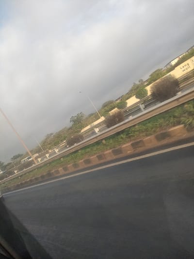 The image captures a diagonally oriented view of a highway section in Murera ward, Kenya, under an overcast sky. The foreground shows a dark asphalt road surface with a visible white lane marker on its right edge. A dark, undefined area occupies the lower-left corner, suggestive of an interior reflection.

A silver-colored metal guardrail runs horizontally across the mid-ground, separating the immediate roadway from an adjacent strip of green grass and a row of dark rectangular concrete blocks. Beyond this, a dense line of varying vegetation is visible, including numerous green trees and several distinct, spherical, brownish bushes.

In the background, a light-colored concrete wall or structure is visible, extending horizontally. Partial text is discernible on this wall, including the word "Wall" towards the upper-right. Two slender, light-colored poles, consistent with streetlights, rise from beyond the guardrail into the sky; one is positioned centrally, and another further to the left. Faint outlines of distant vehicles are visible on a parallel or connecting road in the far left background. No individuals are depicted in the scene.