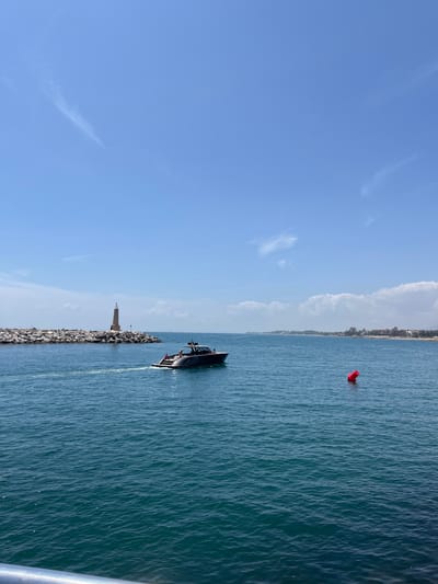 A boat is sailing in the sea toward a breakwater with a light on it. The scene appears to be off the coast of Nueva Andalucía, Spain, identifiable by the red buoy and the coastline in the background. The sky is blue with wisps of clouds.