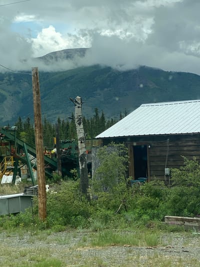 This media file depicts a rural, mountainous scene in Haines Junction, Canada. The focal point is a rustic wooden building with a white corrugated metal roof. In front of the building, there is a large metal structure that appears to be part of an industrial or recreational lift system, possibly a ski lift or a conveyor. A dead tree trunk, stripped of its bark, is positioned vertically within the frame, with a lighter-colored, possibly birch, top portion.
The surrounding environment is lush and green, with dense trees and vegetation. The background is dominated by a large, mist-shrouded mountain range, indicating a natural, outdoor setting. The weather appears overcast, with significant cloud cover and a hint of fog or mist obscuring the mountain peaks, suggesting a cool, potentially damp day. The terrain in the foreground is rocky and covered with grass, typical of a wilderness area.
There are no visible people in the image, and the overall impression is one of a remote, perhaps abandoned, or seldom-used location. The wooden building has a dark, open doorway and a window with a dark interior. Electrical conduits and a junction box are visible on the exterior wall of the building. The metal lift structure has various components, including beams, supports, and what looks like machinery or equipment. A wooden pole with wires runs diagonally across the lower portion of the image.
No visible text is present in the image. The scene evokes a sense of wilderness and solitude, with the imposing mountains and the utilitarian structures hinting at a location with a history of human activity within a wild landscape. The time of day is difficult to ascertain due to the overcast conditions, but it appears to be daytime.