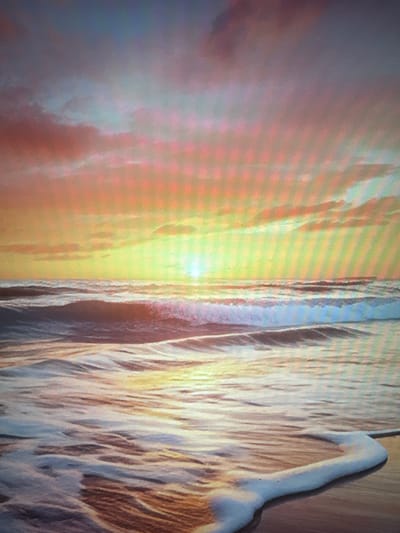The image depicts a serene ocean sunset. The sky is a canvas of warm colors, with hues of orange, pink, and yellow blending into a soft blue at the top. Wispy clouds are scattered across the sky, catching the light of the setting sun. The ocean is calm, with gentle waves rolling towards the shore. The water reflects the colors of the sky, creating a shimmering effect. A line of white foam marks the edge of the water where it meets the wet sand. The scene is tranquil and picturesque, evoking a sense of peace and natural beauty. The image does not contain any people, specific objects, or actions, and therefore cannot be associated with any particular location in Nizhniy Kuranakh, Russia.