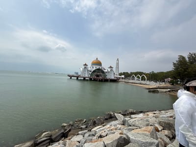 The image depicts a mosque, seemingly floating on water, with a golden dome and a tall minaret. It's connected to the shoreline by a pier-like structure. Arched structures are seen along the shore behind the mosque. The water is calm and reflects the cloudy sky. Rocks line the foreground, and a person in white stands on the right side of the frame.