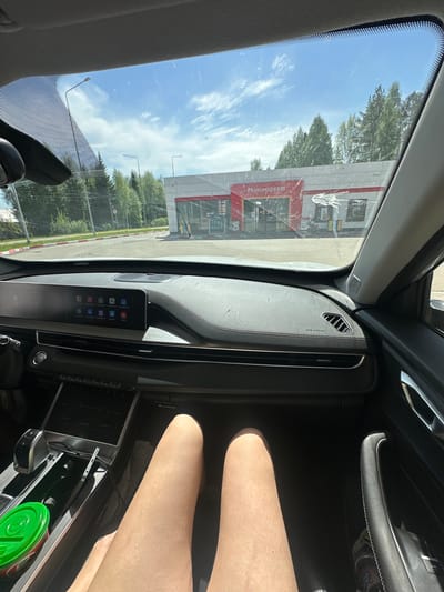 The image shows an interior view from inside a car, looking out the windshield toward a minimarket. The market is a single-story building with a red roof and signage in Russian. Outside, there are trees and a parking area. Inside the car, a dashboard with a screen displaying app icons is visible. A person's legs are visible in the lower right of the frame. A green cup is in the center console.
The location is Uva, Russia.