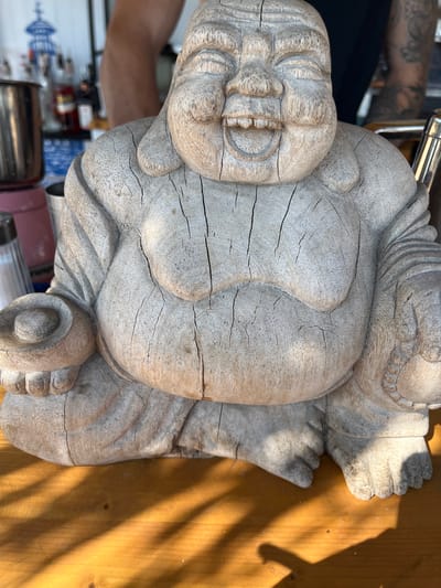 The media file is a close-up, slightly angled shot of a weathered, wooden statue of a smiling Buddha. The statue is carved with visible cracks and grain, giving it an aged appearance. It is positioned on a wooden surface, possibly a table or floor, which is lit by dappled sunlight casting shadows from an unseen source, suggesting it is daytime. The Buddha statue is the primary subject, depicted in a seated position with its legs crossed and hands resting on its lap. Its face is jovial, with a wide, toothy grin and closed eyes. The texture of the wood is prominent, with deep lines and a greyish-white patina. In the blurred background, parts of an arm with tattoos are visible, along with indistinct objects and what appears to be a wall with a blue and white patterned design, possibly from a domestic or public setting. The overall mood is serene and contemplative, emphasized by the peaceful expression of the statue and the warm, natural lighting. No visible text is present in the frame. The location context provided is Dyuni, Bulgaria.
