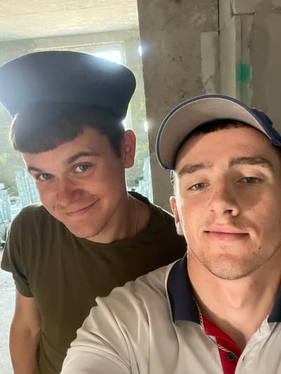 The image shows two young men posing for a selfie indoors, with a background of what appears to be an unfinished or construction site. The man in the foreground is wearing a light-colored polo shirt with navy and red accents and a blue baseball cap. He is looking directly at the camera with a slight smile. The man behind him is wearing a dark olive t-shirt and a dark beret. He is also smiling and looking towards the camera. The environment is dimly lit with some natural light filtering in from a window or opening in the background. The walls appear to be concrete or unfinished plaster, and there are some stacked items, possibly building materials, visible behind the man in the beret. The location is identified as Аснежыцкі сельскі Савет, Belarus.