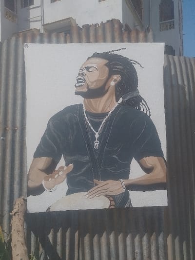 A large, vertically oriented rectangular painting depicting an adult male is affixed to a weathered, vertically corrugated metal fence. The male subject, possessing dark skin, dreadlocks, a beard, and a goatee, is depicted with his eyes closed, mouth open, and head tilted slightly upward and to the left, conveying an expression of intense emotion or vocalization. He wears a dark, short-sleeved shirt, a prominent silver-colored chain necklace with a distinctive pendant, and a white bracelet on his right wrist. His hands are positioned over a light-colored djembe-style drum, with his left hand slightly raised and his right hand resting on the drumhead, indicating active drumming. Behind and above the corrugated fence, a multi-story, light-colored building with visible windows and rooflines is present. A segment of a wooden post is visible in the lower-left foreground, partially obscured by the fence. The sky above is clear. This scene is located in Bamburi ward, Kenya.