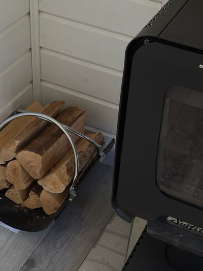 A modern, black wood-burning stove sits in the corner of a room. The stove is positioned on a black base and features a glass door, through which the interior, containing ashes and embers, is visible. A silver metal log holder, filled with several pieces of cut firewood, rests on the floor to the left of the stove. The wall behind the stove and log holder is constructed of white horizontal planks. The floor is covered with light gray wooden planks. The scene appears to be set indoors, possibly in a residence in the village of Alyoshkino DP, Russia.