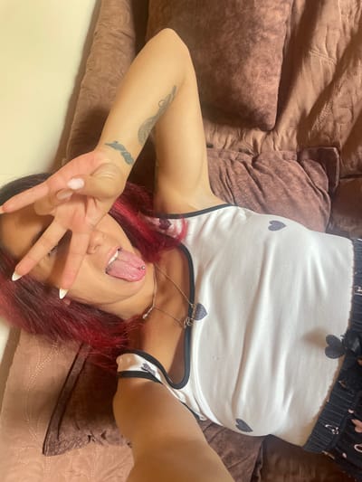 A person with dyed red hair is lying on their side on a brown textured surface. They are wearing a white tank top with small dark heart patterns and black trim, and black shorts with similar heart patterns. The person is holding up two fingers in a peace sign gesture in front of their face, and their tongue, which has a piercing, is sticking out. There are tattoos on the person's arm. The background appears to be a couch or bed. The image does not contain any information to indicate the location is Zheglartsi, Bulgaria.