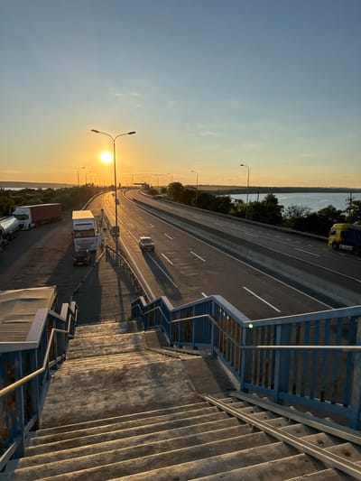 A long shot, vertical, daytime, exterior shot shows a wide highway at sunset. The sun is low in the sky, casting long shadows. The highway has multiple lanes and traffic, including trucks, cars, and a yellow truck. There is a body of water to the right and a landscape of trees and fields. In the foreground, there are concrete stairs with blue railings. The scene appears to be in Yelyzavetivka, Ukraine.