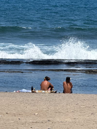 Two individuals are seated on a sandy beach in Playa De Las Américas, Spain, facing the ocean. A wave is crashing against dark, rocky formations in the water. In the foreground, a skateboard, a water bottle, and some rolled-up towels are placed on the sand. The individuals are wearing casual attire, and one has a cap on their head. The sky is clear and blue, and the ocean is a deep blue with white foam from the waves. The scene suggests a relaxing day at the beach.