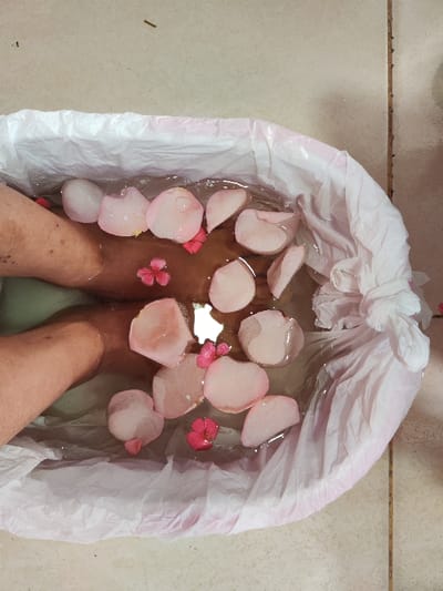 The image shows a top-down view of a foot bath in progress. Two bare feet are submerged in clear water contained within a white, crinkled plastic-lined basin. The water has a slight brownish or reddish tint, possibly from infused elements or light reflection. Floating on the surface of the water are numerous light pink rose petals of varying sizes, along with several smaller, brighter pink flowers, each with five petals. The feet are positioned side by side, with the ankles visible above the water line. The skin on the feet appears light to medium toned. The basin itself is an oval or round shape, and the white plastic liner is crinkled and extends up the sides, giving it a somewhat disposable or protective appearance. The setting appears to be indoors, as indicated by the light-colored tiled floor beneath the basin. In the upper left corner of the image, a small, dark, stick-like object is visible on the tiled floor. The overall impression is one of relaxation and pampering.