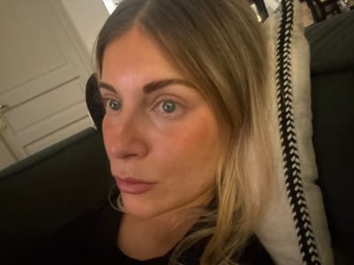 The image captures an adult female from the chest up, positioned indoors within the city of Rueil-Malmaison, France. She has light blonde hair, light blue-grey eyes, and defined eyebrows. Her complexion is fair with a visible flush on her cheeks, and her lips are full, colored with a light rosy-brown tone. She wears a dark-colored, likely black, garment on her upper body. Her gaze is directed off-camera towards the left of the frame, slightly upwards. She appears to be reclining against a dark piece of furniture, with a light-colored decorative pillow, featuring a prominent black braided vertical pattern, positioned to her right. A light-colored, panelled door with a metallic handle is partially visible in the upper left background. The overall lighting suggests an indoor, possibly evening, setting.