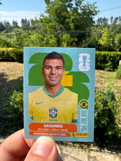This media file shows a close-up, slightly angled view of a person's hand holding a collectible soccer sticker. The sticker features a portrait of a soccer player, identified by the text as "CASEMIRO," wearing a yellow Brazilian national team jersey with the "CBF" logo. The player's name is prominently displayed, along with his birthdate (23-2-1992), height (1.85 m), weight (84 kg), and current club (MANCHESTER UNITED FC (ENG)). The sticker also includes the "FIFA" and "PANINI" logos, indicating its origin and licensing.
The background of the image is blurred, but it appears to be an outdoor setting with green foliage, suggesting a daytime scene with natural light. The presence of trees and grass hints at a park or garden. The sunlight appears bright, casting shadows and illuminating the scene. The hand holding the sticker is in sharp focus, with visible skin texture and fingernails. The sticker itself is colorful, with a light blue background and vibrant graphics. The overall impression is that of someone showcasing a prized soccer collectible outdoors.