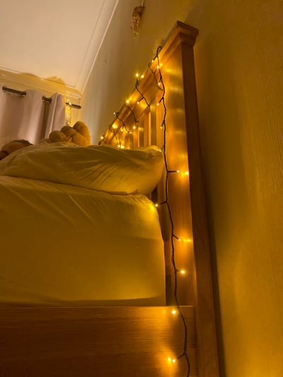 The image shows a wooden bed frame with a string of fairy lights draped across it. The bed is made with white sheets and a pillow. The bed frame is located on the left side of the image. There is a person lying in the bed, but only their head and shoulders are visible. The lights are illuminating the bed frame, and the room is dimly lit. The image was taken at latitude: 51.604, longitude: -1.139, which is located in Wallingford, United Kingdom. The image is taken from a low angle, looking up at the bed frame. The image is in focus and well-lit. The colors are warm and inviting. The image evokes a sense of peace and tranquility.
