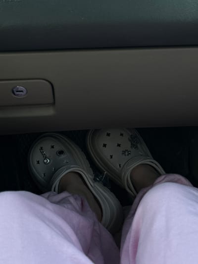 In an indoor environment, possibly a vehicle, a person's legs are visible. The person is wearing light-colored slip-on shoes with decorative charms. The shoes are positioned on a dark floor mat. Above the shoes, a dark gray compartment with a keyhole is visible. The person's lower legs are covered by light pink fabric, likely pants or a skirt. The scene does not provide any specific visual cues to determine the exact location in the city of Töretam, Kazakhstan.