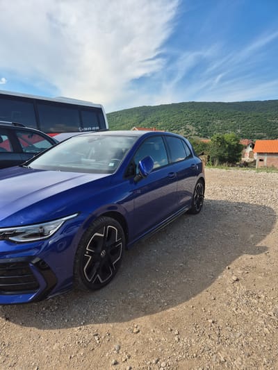 A blue Volkswagen Golf R is parked on a gravel lot. The car is in the foreground, angled towards the viewer's left. Behind the car, a white bus is visible, and further back, a green, tree-covered hill rises under a blue sky with wispy clouds. Several buildings with orange tiled roofs are scattered across the hillside. The scene appears to be in a rural or semi-rural area, possibly in Dimitrovgrad Municipality, Serbia, given the presence of the bus with a Bulgarian phone number (+359 879 455 655). No people are clearly visible in the image, though a reflection in the car's windshield suggests there might be occupants. The lighting indicates it is daytime, with shadows cast by the car and surrounding objects.