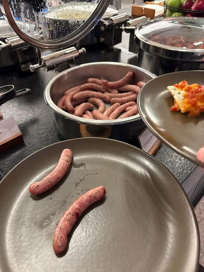 This media file depicts a close-up view of a breakfast buffet setting in Callao, Peru. The primary subjects are a selection of raw sausages in a metal bowl and two partially cooked sausages on a dark gray plate. The sausages appear to be a light pinkish-brown color. In the background, a silver chafing dish with a lid is visible, containing what looks like a baked dish topped with cheese and red ingredients, possibly a casserole or gratin. Another plate with a portion of this baked dish is being held by a person's hand, which is partially visible at the top of the frame. The overall scene suggests a meal being served, likely breakfast, given the types of food shown. The lighting is artificial, indicative of an indoor setting, and there are no obvious cues about the time of day or weather. The objects are arranged on a dark countertop, with some reflections on the metallic surfaces. No visible text is discernible in the image. The focus is on the food items, with an emphasis on the preparation or serving process.