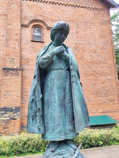A full-shot, eye-level, outdoor, overcast photograph shows a bronze statue of a young woman in traditional Russian dress standing in front of a brick building. The statue is weathered and has a green patina. The woman wears a fur-trimmed hat and a long, ornate robe with a patterned skirt. Her hands are clasped at her chest. The building behind her is made of red brick and has a small arched window. There are green bushes and a green metal shed at the base of the building. The ground in front of the statue is paved with brick. The scene appears to be located in Chelganovo, Russia.