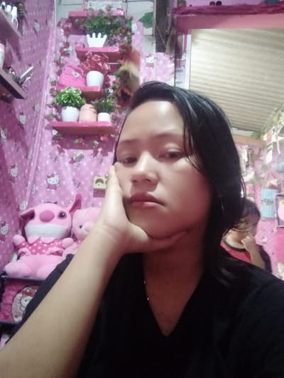 A person with short black hair rests their head on their hand, gazing directly forward with a solemn expression. The backdrop features a bright pink room adorned with Hello Kitty-themed wallpaper, plush toys, and tiered shelves showcasing various potted plants. A second person is partially visible in the background, adding depth to the scene. The overall aesthetic is playful and feminine, contrasting with the individual's serious demeanor.