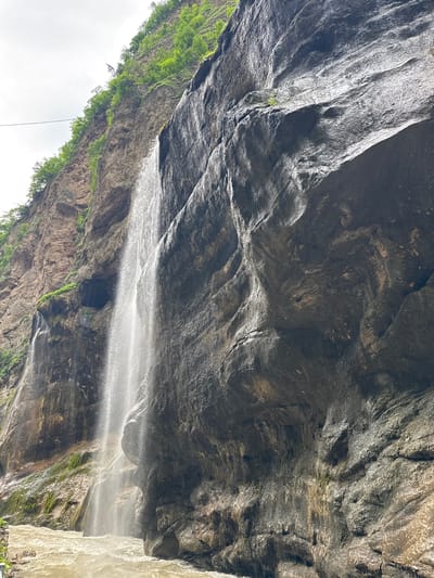 The image depicts a view of the Khushto-Syrt waterfall in Russia. A cascading waterfall, appearing as a thin stream of water, descends from a high cliff face on the left side of the frame. The cliff is a mix of rough, rocky textures and patches of green vegetation clinging to its upper ledges. The rock face is dark and wet, reflecting the light. To the right of the waterfall, a massive, dark, and uneven rock formation dominates the scene, with intricate patterns and textures on its surface. At the base of the waterfall, a turbulent, muddy river flows, with visible ripples and waves. The sky above is overcast and light. The overall impression is one of a natural, somewhat rugged, and powerful landscape.