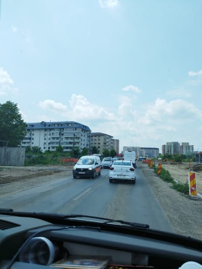 The image shows a view from inside a vehicle looking out at a street scene in Bragadiru, Romania. The street is a two-lane asphalt road with traffic. In the foreground, the dashboard and windshield wipers of the vehicle are visible.
On the road, several cars are present. A white van with the license plate B 421ZHH is in the middle lane. Ahead of the van is a white Dacia car with the license plate B 94 YAR. Further ahead, there are other vehicles, including another white van and a white car.
The environment indicates construction is taking place. The right side of the road is lined with construction barriers, including yellow and red striped traffic cones and other orange construction equipment. The left side of the road features a tall, multi-story residential building with a dark roof and numerous windows. Adjacent to this building is a concrete wall. Beyond the buildings, the sky is partly cloudy and blue. The overall impression is a suburban or urban area undergoing road work.