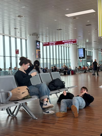 The image shows the interior of an airport, specifically Orio al Serio International Airport in Orio al Serio, Italy. A young woman with dark hair pulled back and wearing a black top sits on an airport seat, with a beige backpack next to her. Another person, also wearing jeans, sits on the seat next to her. Further along, a young man is on the floor near the seats, partially lying down and looking toward the camera. Several people are seated or standing in the background waiting for their flights. Signage in the airport includes gate information (A3-A16), as well as directional signage in both Italian and English ("RITIRO BAGAGLI-USCITA / Baggage claim-Exit"). Large windows provide natural light into the airport space.
