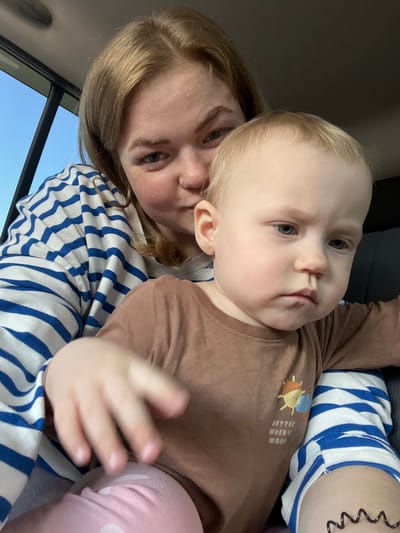 This media file is a selfie taken from a low angle within a vehicle. The primary subjects are a woman and a baby. The woman, with blonde hair and wearing a blue and white striped shirt, is positioned on the left side of the frame, her face turned towards the camera. She has a nose ring and appears to be looking at the camera.

The baby, with fair skin and light blonde hair, is being held by the woman and occupies the center and right portion of the frame. The baby is looking slightly to the side, with a neutral expression. The baby is wearing a brown long-sleeved shirt with a small graphic of a character and the text "BETTER WHEN WARM" printed on it. The baby also appears to be wearing pink leggings with white polka dots. The woman is wearing a blue and white striped long-sleeved shirt or sweater.

The setting appears to be the interior of a car, indicated by the car seat visible behind the woman and the sky visible through what seems to be a car window on the left side of the frame. The sky is a clear, bright blue, suggesting daytime and fair weather. The image is well-lit, likely by natural light coming from the window. There are no strong indications of a specific time of day beyond it being daylight. The overall mood conveyed is casual and affectionate, typical of a parent and child sharing a moment. The crops provided highlight the faces of the subjects, the text on the baby's shirt, and the clothing details. The provided location context is Krilovo, Russia.