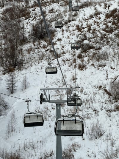 The image displays an operational ski chairlift system ascending a snow-covered mountain slope in Novoabzakovo, Russia. A prominent grey metal chairlift pylon, marked with the numeral '6', stands in the lower-central portion of the frame. Multiple grey chairlift chairs are suspended from a thick steel cable, extending diagonally upwards and downwards across the scene. One chair, positioned immediately above and to the right of the pylon, bears the number '79' and the text "ABZAKOVO" faintly visible on its backrest. Higher on the slope, another chair is occupied by two indistinct individuals, one of whom is partially visible wearing a red garment. The mountain terrain is covered by a thin, patchy layer of snow, revealing dry, brown vegetation and sparse, bare deciduous trees. Several young evergreen trees are also visible. The lighting is soft and diffuse, indicating an overcast sky. Faint tracks or trails are present in the snow on the lower left portion of the slope.