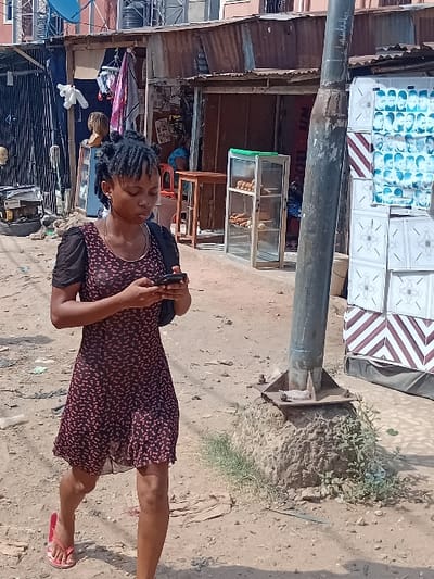 An outdoor scene in Ifite Awka, Nigeria, depicts a young adult female walking along a dirt path on a bright day. The female, wearing a short-sleeved, dark red patterned dress and red flip-flops, has her hair styled in multiple dark buns or dreadlocks. She is actively engaged with a black smartphone, holding it with both hands and looking down at its screen. A dark shoulder bag hangs from her left shoulder.
To her right, a tall, dark grey metal pole with a concrete base stands on the dirt. Adjacent to the pole is a display constructed from stacked white rectangular panels with brown geometric patterns, featuring a poster with multiple small headshot images, likely an advertisement for a barbershop or salon.
In the background, a row of informal commercial structures with corrugated metal roofs and walls lines the path. The shop immediately behind the female features a black accordion-style metal gate, a white plush toy hanging, various fabrics, a wig mannequin head, and a generator. Further right, another shop interior is visible, showing a wooden table, a red plastic chair, and a person in blue clothing seated. In front of this shop, a glass display case contains several baked goods. Above the shops, a multi-story building with red-colored walls and barred windows is visible, topped by a blue satellite dish. The ground is unpaved, consisting of packed dirt with scattered debris and some small green plants near the pole's base.