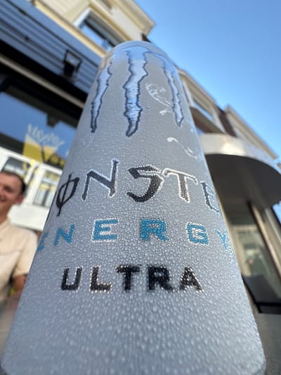 A close-up, low-angle shot focuses on a frosty can of Monster Energy Ultra, its surface covered in condensation. The can is tilted, with the logo and text "MONSTER ENERGY ULTRA" visible. In the background, out of focus, is a man with short brown hair wearing a light-colored shirt, smiling. Behind him, a building with a dark horizontal siding and windows is visible, along with a bright blue sky. The scene appears to be outdoors, possibly on a patio or terrace, in the city of Deventer, Netherlands.