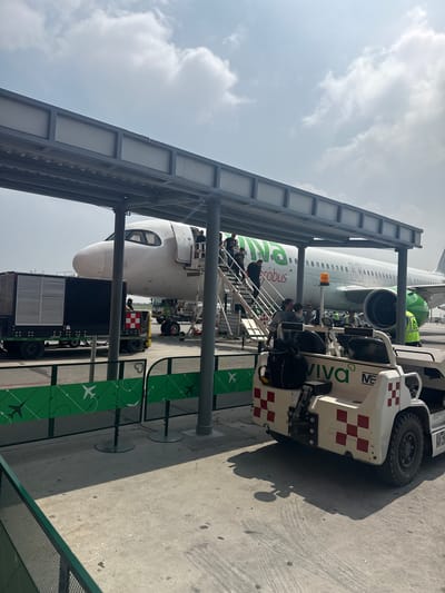 The image shows a Viva Aerobus airplane being boarded at an airport, likely in Apodaca, Mexico, based on the airline's presence there. Passengers are walking up the stairs to the aircraft. A baggage handling vehicle is parked nearby. The sky is overcast. The scene is viewed from within a boarding bridge structure. A ground services vehicle is visible with "Viva Aerobus" branding.