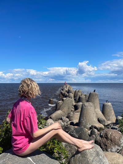 A young woman with blonde, curly hair sits on large rocks facing away from the camera, looking out at a body of water. She is wearing a bright pink t-shirt and her legs are visible. The rocks are part of a breakwater that extends into the water, made of large, concrete tetrapods. A red navigation beacon is visible in the distance at the end of the breakwater. The sky is blue with scattered white clouds. The scene is likely on the shore of Lake Beloye in Belozersk, Russia.