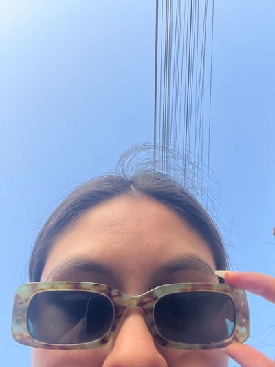 The image is a close-up, low-angle shot of a person wearing tortoise-shell sunglasses against a bright blue sky. The person's forehead and eyebrows are visible above the sunglasses, with strands of dark hair framing their face. Several thin, dark wires hang vertically in the upper portion of the frame, suggesting an outdoor urban environment. The person's right hand is visible on the right side of the frame, adjusting the sunglasses. The image appears to be a selfie. The location is specified as San Borja, Peru.