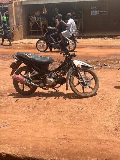 The image shows a scene in Rigasa, Nigeria. A dirt road is visible in the foreground. Two motorcycles are present. One motorcycle is in a state of disrepair, with its front wheel removed and placed beside the motorcycle. Another motorcycle is parked in front of a building, where several people are gathered. One man wearing a white shirt and a cap is leaning over the motorcycle, possibly examining or repairing it. The building appears to be a workshop or storefront.