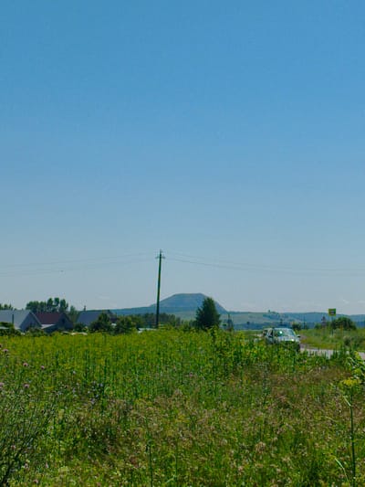 The image depicts a rural landscape under a clear blue sky. In the foreground, a dense field of green grass and wildflowers stretches across the frame. In the mid-ground, several houses with pitched roofs are visible, nestled amongst trees. A single utility pole with wires stands prominently to the right of the houses. Further in the distance, rolling hills form the horizon, with a large, conical mound dominating the skyline. A paved road can be seen in the right mid-ground, with a white car driving on it. Two road signs are also present near the road. The scene appears to be a sunny day in a quiet, possibly remote, location. Based on the provided information, this scene is located in Vostok, Russia.