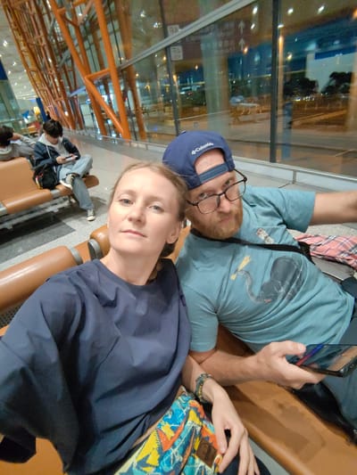 Two individuals, a woman and a man, are sitting on a tan-colored bench. The woman is wearing a navy blue top, and the man is wearing a light blue t-shirt with an elephant design and a blue baseball cap with a visible brand logo. He is also wearing glasses and holding a smartphone. They appear to be posing for a photograph. In the background, other people are sitting on similar benches, and an orange architectural structure is visible through a glass window. The scene appears to be taking place in an airport, potentially in Shunyi District, China.