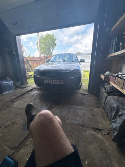 The image shows the interior of a garage in Borodulino, Russia. A dark-colored car is parked partially inside the garage, facing outwards towards a sunny sky and a wooden fence. The garage floor is concrete and appears dusty and cluttered with various items, including a black plastic-wrapped object on the left and shelves filled with storage containers and tools on the right. In the foreground, a person's legs and feet, clad in dark boots, are visible, indicating they are sitting or reclining within the garage. The overall scene suggests a typical residential garage environment in a rural or suburban setting.