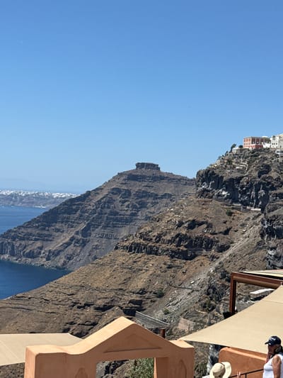 The image depicts a scenic view of the caldera cliffs in Santorini, Greece, likely taken from a vantage point in Fira. The foreground features architectural elements characteristic of the Cycladic style, including an orange-hued building with a distinctive curved opening and a large, light-colored awning. Below the awning, a person wearing a white top and a black baseball cap is partially visible, looking downwards. Another person, wearing a straw hat, is also present, partially obscured.

The middle ground is dominated by the rugged, rocky slopes of the caldera, with evidence of a pathway winding up the side of the cliff. In the distance, a settlement with white-washed buildings is perched on the edge of the caldera, with a prominent red-roofed building among them. The deep blue Aegean Sea stretches out to the horizon, meeting a clear, bright blue sky. A flat-topped mountain or rock formation rises from the sea on the left side of the image, adding to the dramatic landscape. The overall scene captures the breathtaking geological and architectural beauty of Santorini.