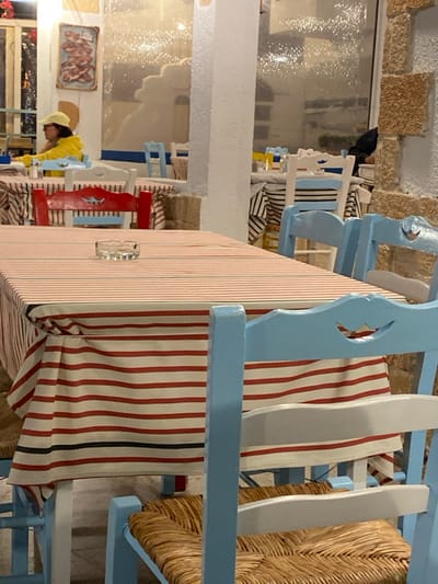 The image depicts the indoor seating area of a restaurant or tavern in Megalochori, Greece. In the immediate foreground, a rectangular table covered with a white tablecloth featuring horizontal red and thin black stripes holds a clear glass ashtray. To the left, a person wearing a yellow jacket and a light yellow baseball cap is seated at another table with a similar striped tablecloth, their left arm resting on the surface. A red wooden chair with a decorative carved backrest is visible in front of this table, which also holds salt and pepper shakers. Numerous light blue wooden chairs with woven straw seats are arranged around multiple tables throughout the scene. Some tables are covered with white tablecloths featuring horizontal blue and black stripes. In the background to the left, a framed picture depicting multiple fish is mounted on a white wall. The central background features a large, textured glass partition or window, reflecting light. To the far right, a wall constructed of rough-hewn, light-colored stone blocks is visible. A dark figure is partially obscured behind a table near this stone wall. The environment is illuminated by artificial light.