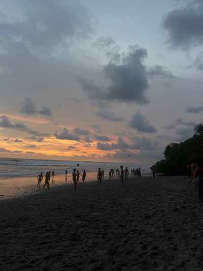 The image shows a group of people playing a game of soccer on a beach in Santa Fe, Costa Rica. The coordinates provided place the scene near Playa Santa Fe. The sky is mostly cloudy, with some sunset colors visible in the distance. The beach is sandy and wet, and the water is visible in the background. There is a line of trees on the right side of the image. The majority of the people in the image are on the beach, playing soccer. There are several figures standing in the water and some on the edge of the tree line. The action in the image is focused on the people playing soccer. They are kicking a ball around and interacting with each other. The scene is set during the evening with a soft light illuminating the scene.