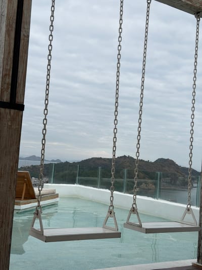 The image shows a view from a rooftop swimming pool area overlooking a scenic landscape, likely in Labuan Bajo, Indonesia, given the presence of islands and a harbor. In the foreground, three white swing seats are suspended from metal chains attached to a wooden beam. The swings are positioned over the shallow, pale blue water of the pool. To the left, a wooden lounge chair with a beige cushion is visible. A white railing with glass panels separates the pool area from the outdoor environment. Beyond the railing, a hillside covered in dry vegetation descends towards a body of water. In the distance, several islands with hilly terrain are scattered across the horizon under a cloudy sky. The overall atmosphere is serene and offers a panoramic view of the natural surroundings and the coast.