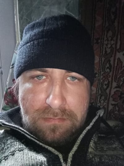 A close-up, medium-shot photograph features a man with a black beanie, light eyes, a fair complexion, and a beard and mustache. His skin appears to have some blemishes. He is wearing a dark-colored jacket with a light-colored trim. Behind him, there is a patterned curtain or tapestry. To the left, there appears to be a window with a sheer curtain. The photograph does not contain enough information to establish its location in Parabel, Russia.